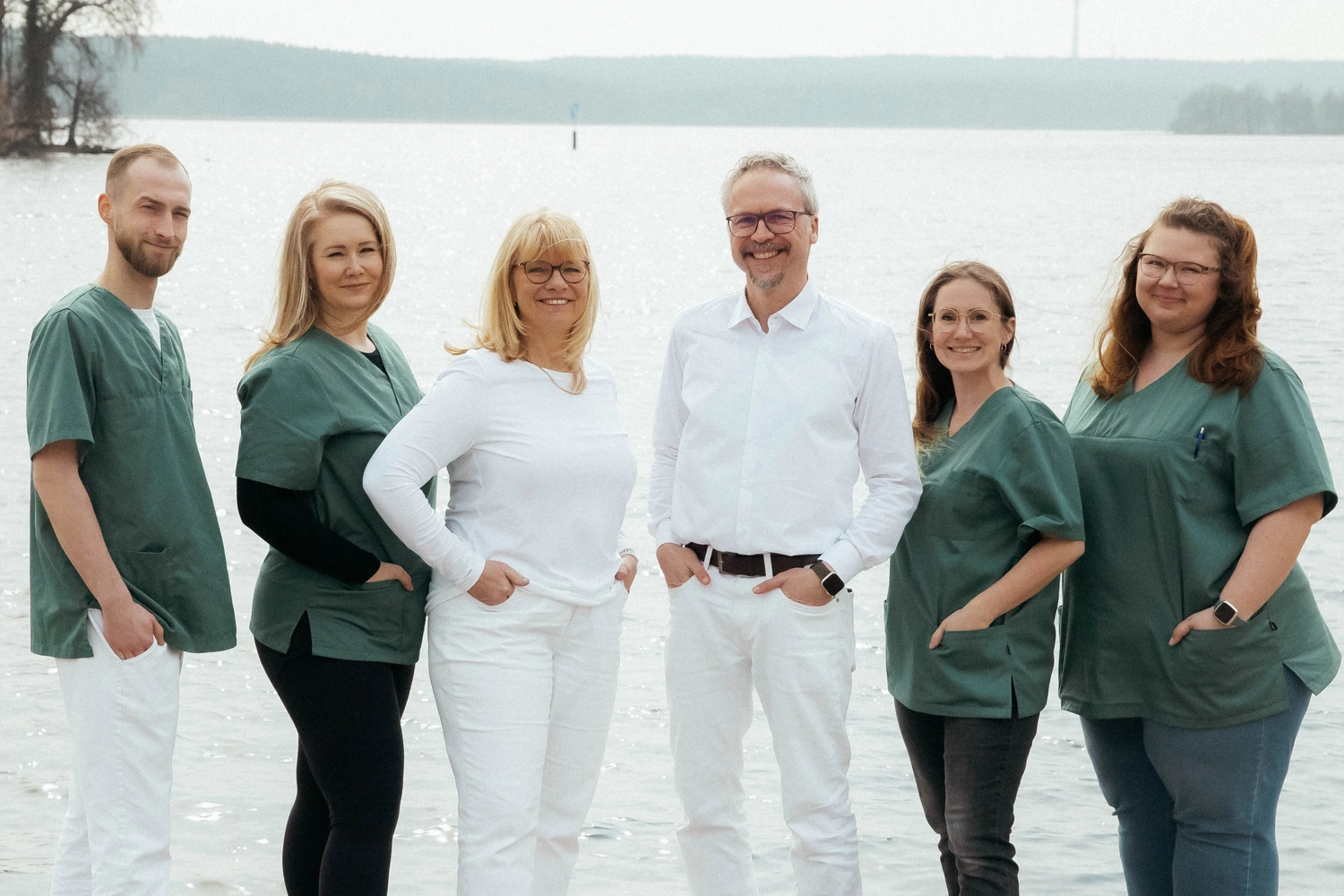 Praxis an der Havel - Innere Medizin, Diagnostik, Prävention und chronische Versorgung - Sechs Personen stehen in einer Reihe am Strand, im Hintergrund sind Wasser und Bäume zu sehen. Drei Frauen und ein Mann tragen dunkelgrüne OP-Hemden, während eine Frau und ein Mann in der Mitte ganz in Weiß gekleidet sind. Alle lächeln in die Kamera.
