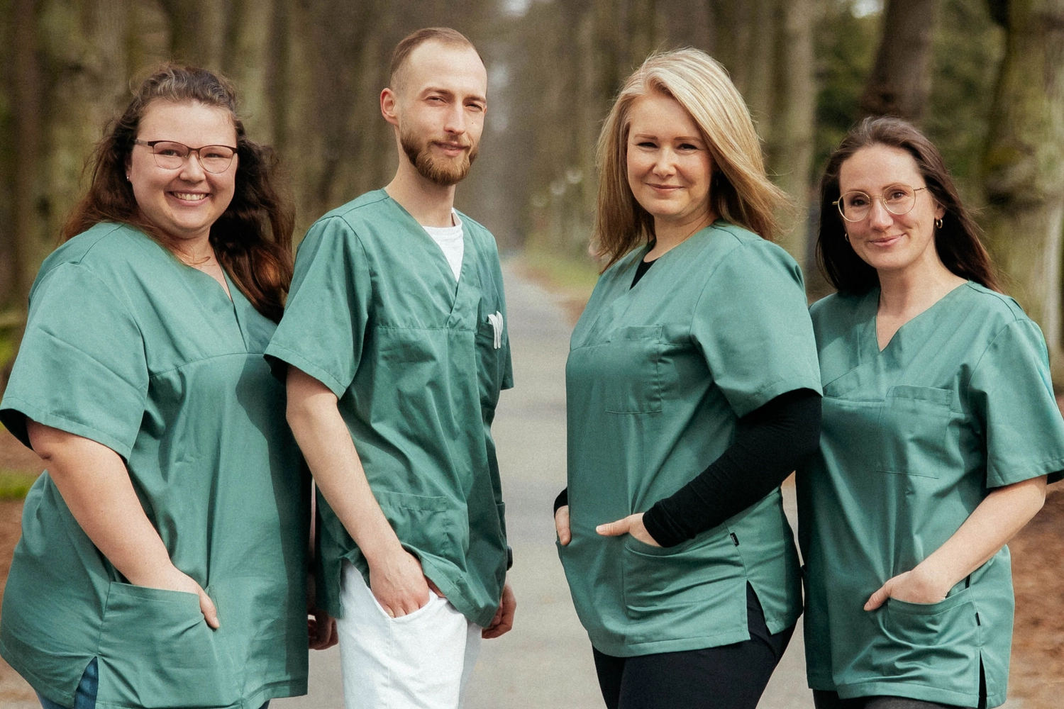 Praxis an der Havel - Innere Medizin, Diagnostik, Prävention und chronische Versorgung - Vier Angehörige der Gesundheitsberufe stehen im Freien nebeneinander auf einem Weg, alle in identischer salbeigrüner OP-Kleidung.