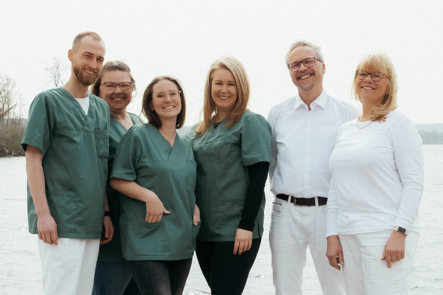 Praxis an der Havel - Innere Medizin, Diagnostik, Prävention und chronische Versorgung - Sechs Personen stehen in einer Reihe am Strand, im Hintergrund sind Wasser und Bäume zu sehen. Drei Frauen und ein Mann tragen dunkelgrüne OP-Hemden, während eine Frau und ein Mann in der Mitte ganz in Weiß gekleidet sind. Alle lächeln in die Kamera.