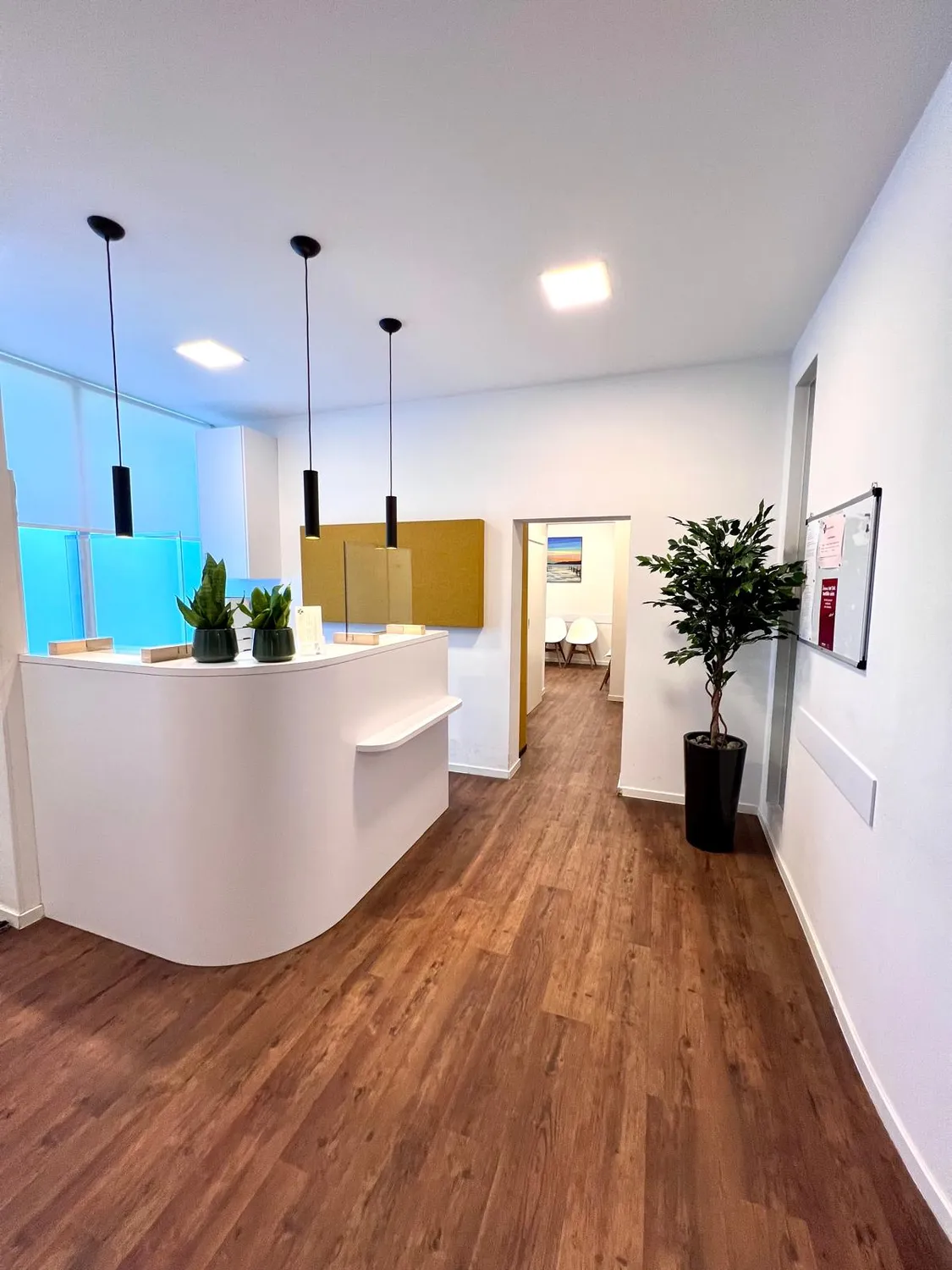Praxis an der Havel - Innere Medizin, Diagnostik, Prävention und chronische Versorgung - Ein hell erleuchteter, moderner Empfangsbereich mit einem geschwungenen weißen Schreibtisch, Holzbodenoptik und drei minimalistischen schwarzen Pendelleuchten. Neben einer weißen Tür, die in einen anderen Raum führt, steht ein Zimmerbaum in einem hohen schwarzen Übertopf.