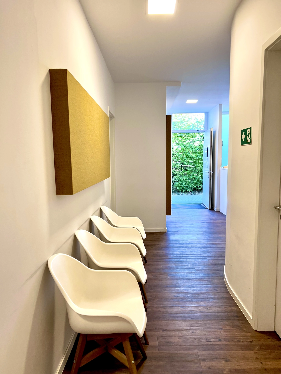 Praxis an der Havel - Innere Medizin, Diagnostik, Prävention und chronische Versorgung - Ein heller, moderner Flur mit einer Reihe weißer, formgepresster Stühle mit Holzbeinen an einer weißen Wand. Über den Stühlen ist eine große, rechteckige, gelbe Akustikwand angebracht, und der Raum öffnet sich zu einem hellen Türbereich mit Blick ins Grüne.