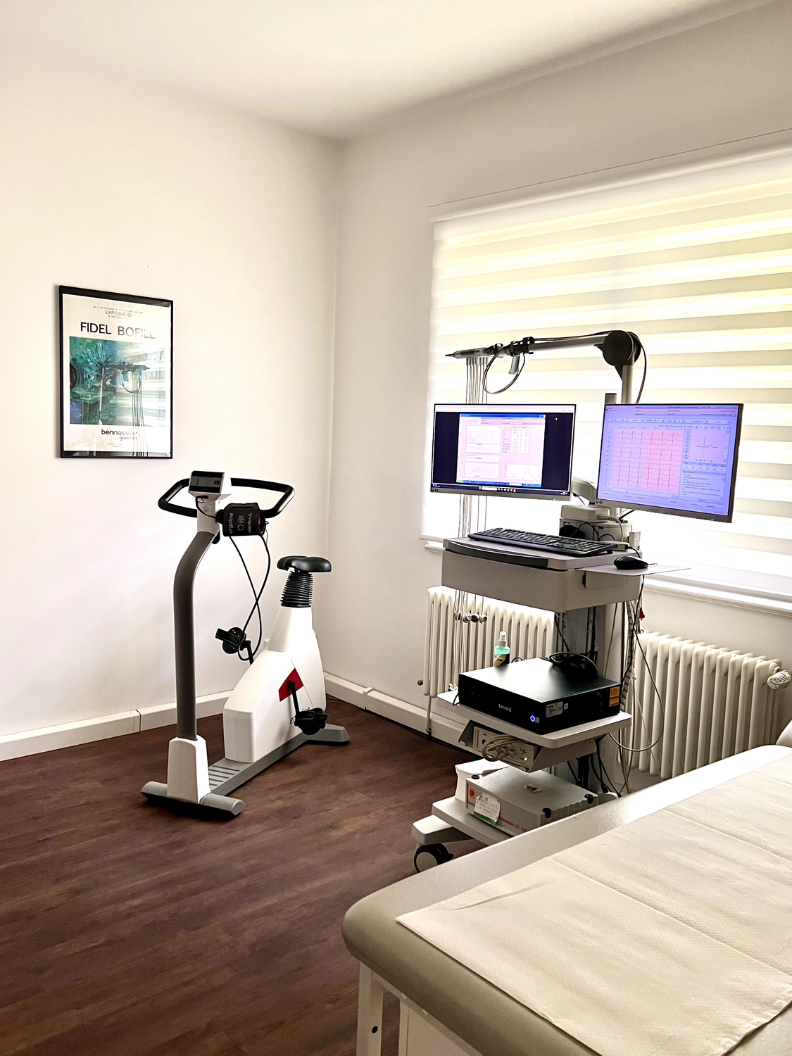 Ein klinischer Untersuchungsraum mit einem medizinischen stationären Fahrradergometer und einer mobilen Workstation mit zwei Monitoren zur Anzeige von Diagnosedaten.