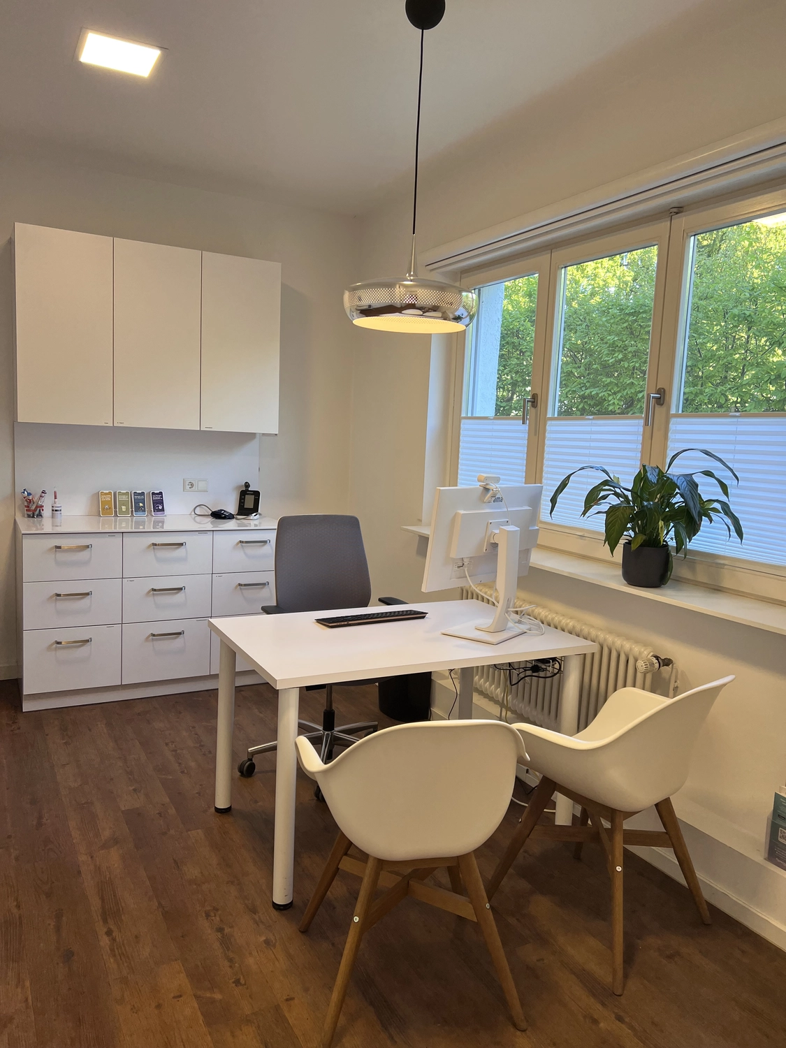 Praxis an der Havel - Innere Medizin, Diagnostik, Prävention und chronische Versorgung - Ein minimalistisches und modernes Homeoffice oder Beratungszimmer mit weißem Schreibtisch, grauem Bürostuhl und zwei weißen Stühlen mit Holzbeinen. An der linken Wand steht ein niedriges Metallregal, und ein großes Fenster mit Plissees lässt viel Tageslicht herein.