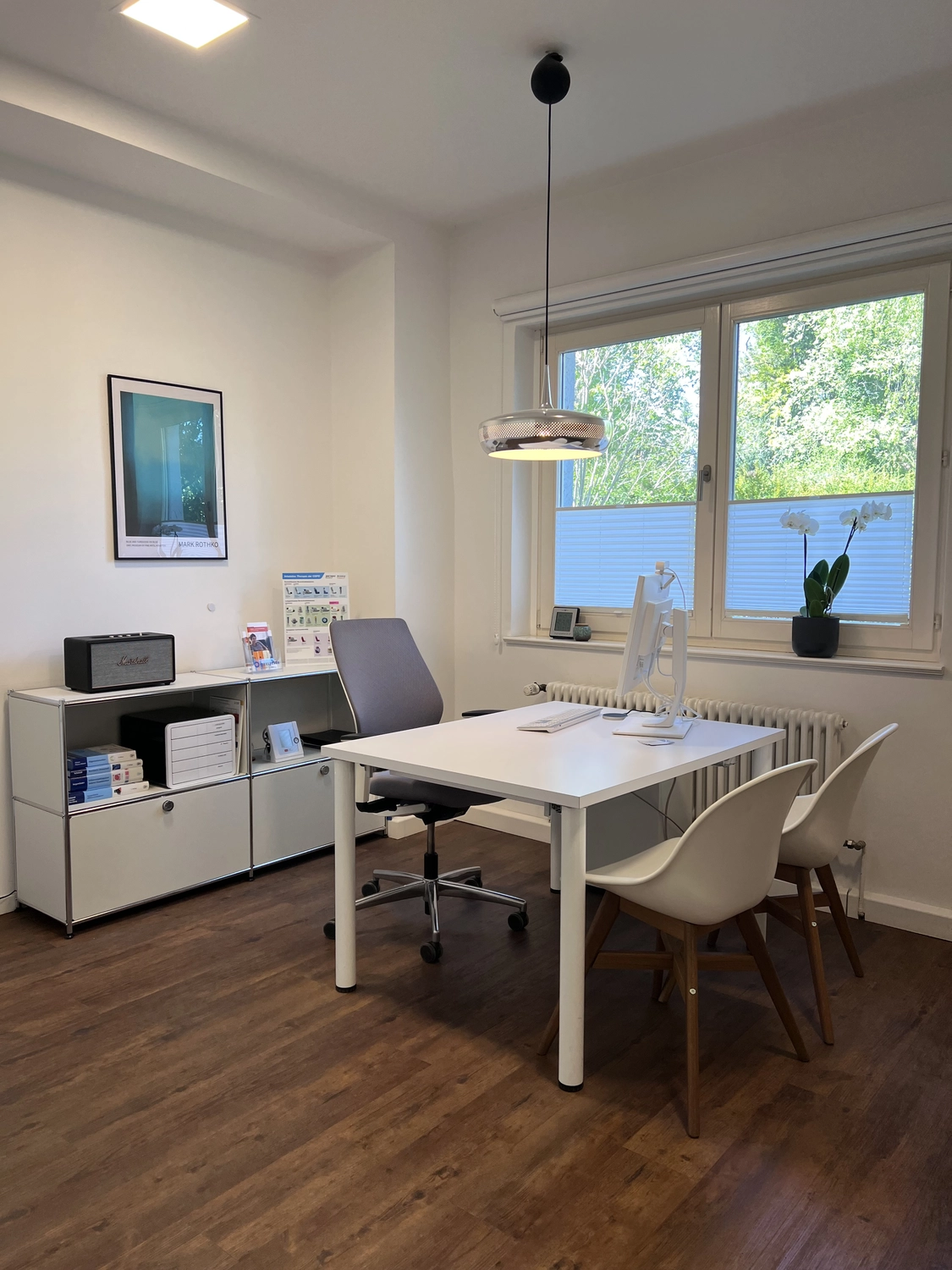 Praxis an der Havel - Innere Medizin, Diagnostik, Prävention und chronische Versorgung - Ein minimalistisches und modernes Homeoffice oder Beratungszimmer mit weißem Schreibtisch, grauem Bürostuhl und zwei weißen Stühlen mit Holzbeinen. An der linken Wand steht ein niedriges Metallregal, und ein großes Fenster mit Plissees lässt viel Tageslicht herein.