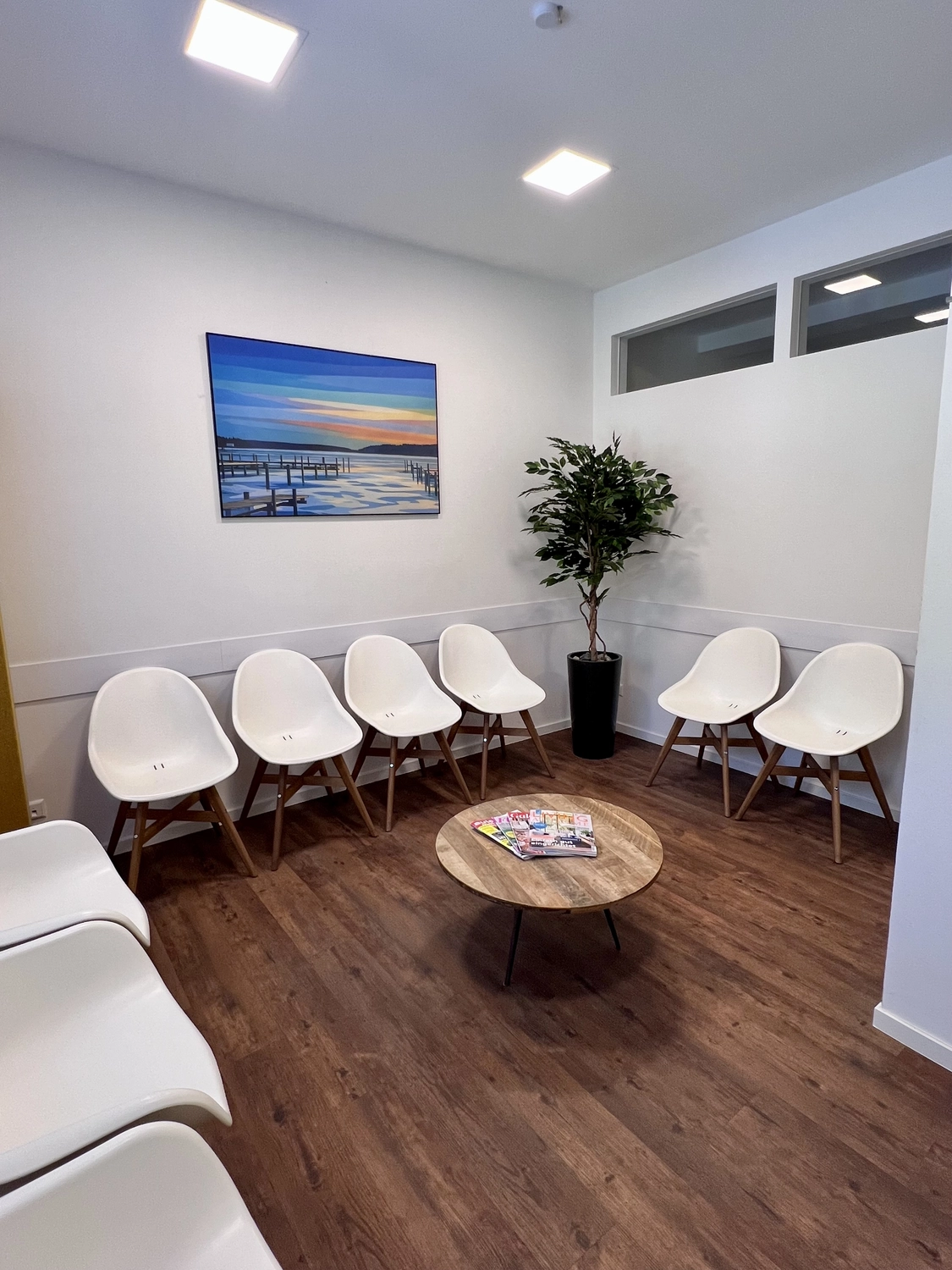 Praxis an der Havel - Innere Medizin, Diagnostik, Prävention und chronische Versorgung - Ein modernes, hell erleuchtetes Wartezimmer mit mehreren weißen, formgepressten Kunststoffstühlen mit Holzbeinen, die an den Wänden stehen, einem niedrigen Holztisch mit Zeitschriften in der Mitte und einer hohen künstlichen Pflanze in der Ecke. An der weißen Wand hängt ein Landschaftsgemälde mit einem Sonnenuntergang über dem Wasser.